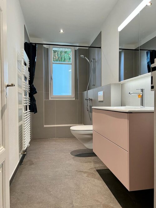 Modernes Badezimmer mit schönen Bodenplatten in dunklem Natursteinoptik und schlichten Wandplatten in grau. Alles in Grossformat und Feinsteinzeug. Das rosa Waschbecken passt sehr gut zu der gesamten Farbauswahl.