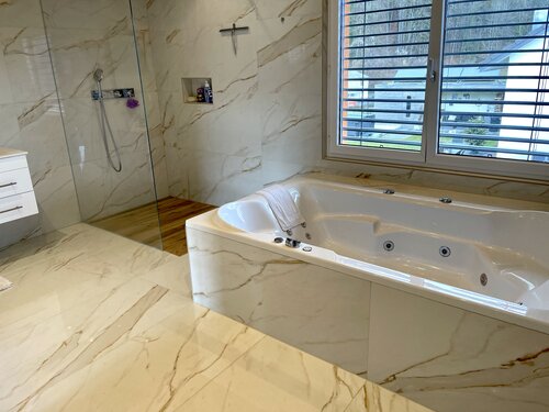 Grosszügiges Badezimmer mit Whirlpool und Bodenebender Dusche. Eine Kombination aus Feinsteinzeug in goldener Marmoroptik und Keramik in Holzoptik am Boden der Dusche bildet ein harmonisches Zusammenspiel.