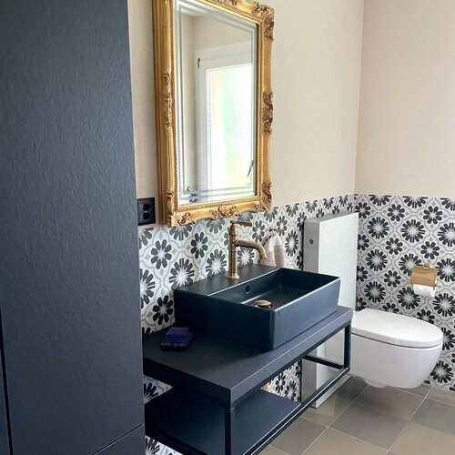 WC mit Schachbrett Boden in braunton und Dekor Wandplatten auf 1.2 Meter Höhe. Das schwarze Waschbecken mit den goldenen Armaturen und dem goldenen Spiegel runden das Bild ab.