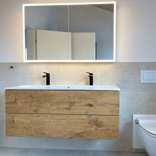 Badezimmer mit Wandplatten welche feine Rillen haben für den besonderen Effekt. Passend dazu schwarze Armaturen, LED Spiegel, Holzmöbel